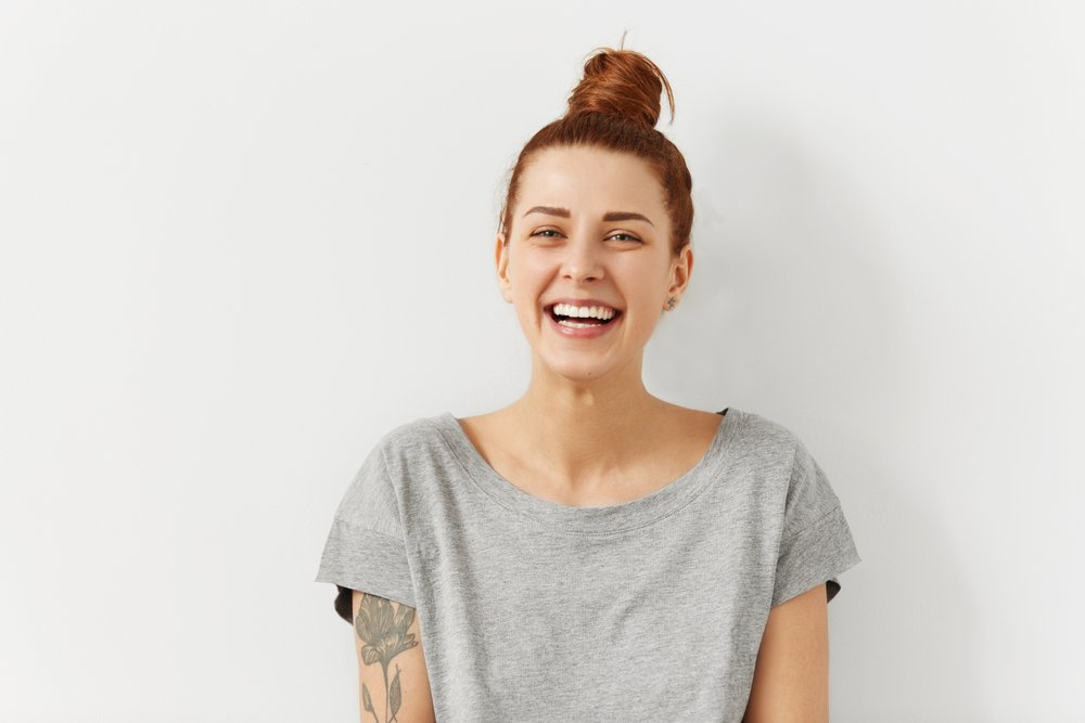 Happy cheerful young woman wearing her red hair in bun rejoicing at positive news or birthday gift, looking at camera with joyful and charming smile. Ginger student girl relaxing indoors after college